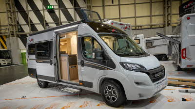 Malibu Van diversity GT skyview 640’s exterior is white with back and gold decals.  Its sliding door is open revealing the kitchen.  There is a step to gain easy access.  The roof light is open.