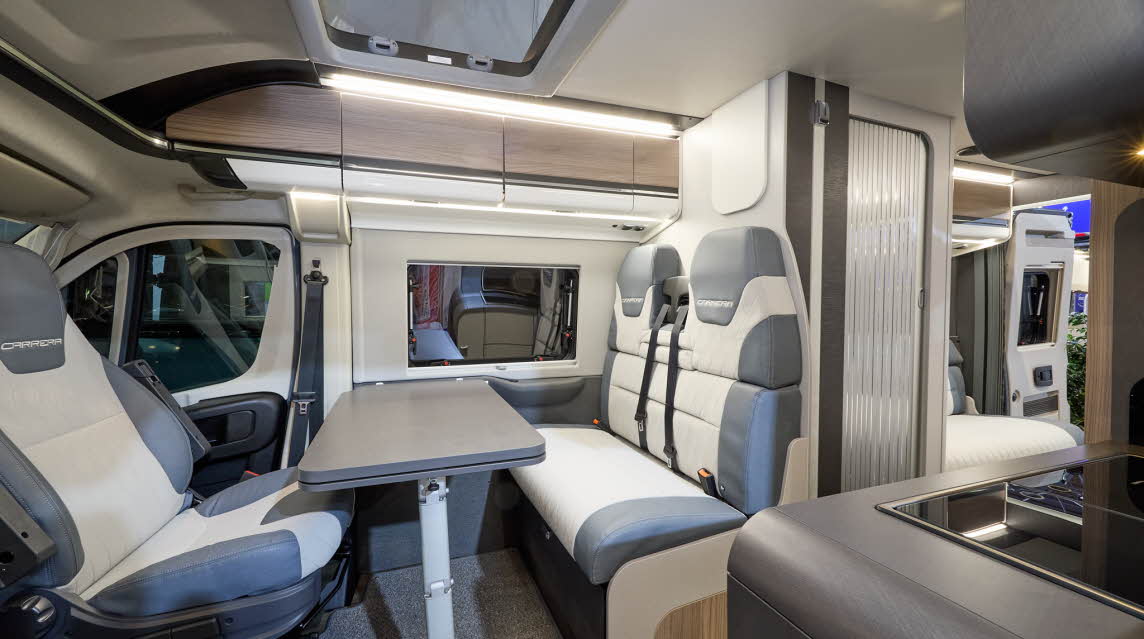 The Swift Carrera 244 has a grey floor and its upholstery is pale grey and blue.  The cabinets are wood with white handles.  The rising roof is open. Towards the rear there is lounge and one of its rear doors are open.