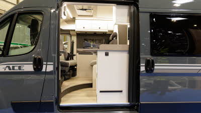 The Ace Motorhomes 1200 RL is grey with white decals, its sliding door is open revealing a pale wooden floor with matching cabinets.