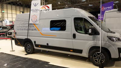 The WildAx Motorhomes Solaris XL is grey with black and orange decals.  There is a flag with its base beside the rear wheel.