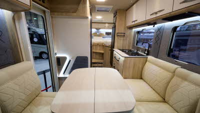 The Eura Mobil Xtura XT 686 EF has cream and wooden cabinets.  The upholstery is cream leather.  There is a cream table in front of the sofas.  There are two steps to the rear fixed beds.