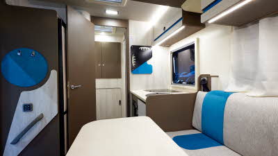 Rimor Kilig 67 has a cream interior with dark wooden doors.  There are blue accents in the upholstery, overhead lockers and décor. 