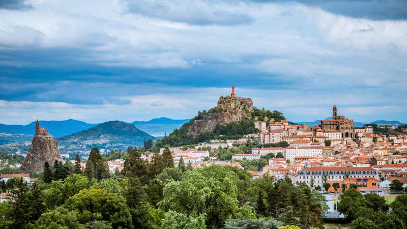 An elevated view looking out to the city of Le Puy, France. Trees of various shades of green leading to the buildings on the hills under a blue sky with white clouds 