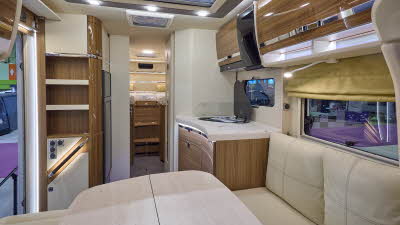The Eura Mobil Contura CT 766 EB has grey carpets and shiny wooden cabinets.  The upholstery is cream leather.  There is a large cream table in the front.  There are two steps to the rear fixed bed.