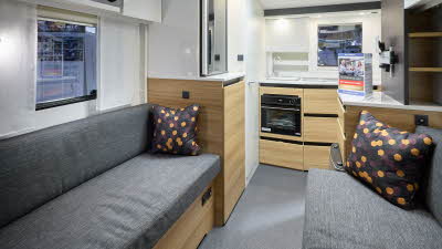 The interior of the Adria Action 361 LT has white walls.  The furniture is mid wood.  There are two sofas with grey upholstery and a cushion with a hexagon pattern on each.  The kitchen is at the rear with a full oven under the sink with a mixer tap.