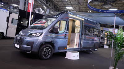 The Auto-Trail V-Line 635 Sport is grey with red decals.  There is a show offer on its side in red.  There is a white step by the open sliding door which reveals the interior.  