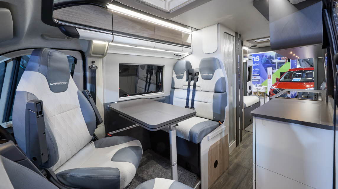 The Swift Carrera 184 has a wooden floor and its upholstery is pale grey and blue.  The cabinets are  wood with white handles.  Towards the rear there is lounge and both rear doors are open.