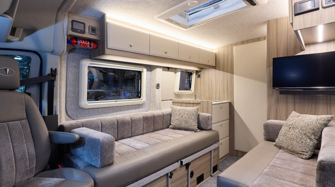 Vantage ORA F-Line has grey carpet, with white and wooden cabinets.  The upholstery is brown leather with a matching pattern insets.  There is a rooflight.  The cream internal door is closed.  There is a TV above a sofa.