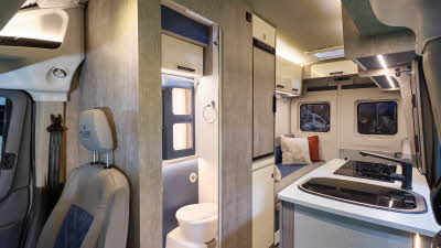WildAx Motorhomes Meteor has wooden cabinets, its upholstery is pale leather with a blue inset.  The washroom door is open revealing the toilet.  There is a rear lounge.