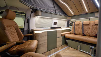 The Sherwood Campers Ltd Loxley Edition Conversion has wooden parquet-style flooring and its upholstery is brown leather.  Its furniture is wooden which is curved.  There are dark green inserts.  The rising roof is open, there's strip lighting.