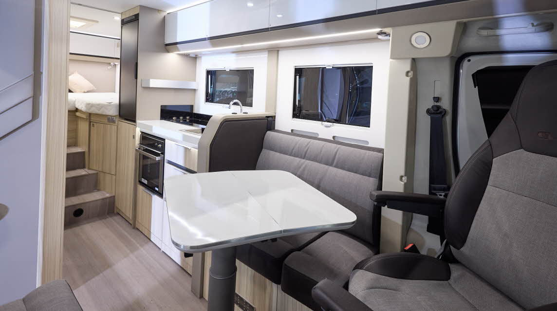 The Adria Compact Supreme has a wood style floor, with pale wood cabinets and white overhead lockers.  The upholstery is two tone with dark edging.  There is a white table which can be rotated.  There are three steps to the fixed raised rear bed.