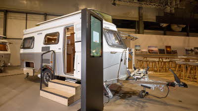 The classically styled Eriba Touring 540 Legend has a two tone layout in silver and white.  Its pop top roof is open as is its entrance door.  There are two wooden steps to gain easy access.