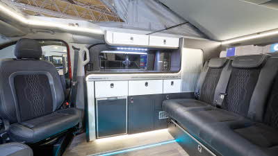 The Swift Monza has wooden flooring with black upholstery. The cabinets are white and dark blue. The pop top roof, along with its canvas, is open.