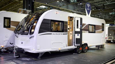 The Bailey Alicanto Grande Alvor has a white exterior with a dark black and brown decal including lighter stripes,  The front door is open and there is a Wi-Fi teardrop sign on its roof.