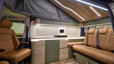 The Sherwood Campers Ltd Loxley Edition Conversion has wooden parquet-style flooring and its upholstery is brown leather. Its furniture is wooden which is curved. There are dark green inserts. The rising roof is open, there's strip lighting.