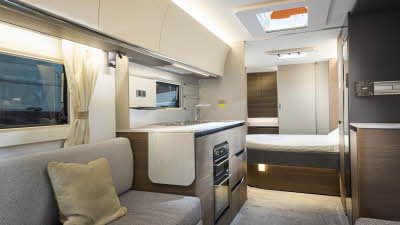 Interior of the Adria Alpina Mississippi with grey sofas, cosy cushions and wood accents, illuminated by natural light through windows and skylights.  There is a fixed island bed and the door at the rear leads to the washroom.