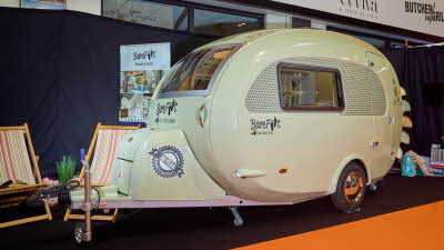 Barefoot Caravan Country Living x Barefoot’s exterior is pale green.  There are two stripy deck chairs at the hitch end with a grass plant 