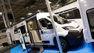 The Sun Living V Series 65 GX has a white exterior with black trim.  Its exterior door is open and there are two black steps to gain easy access.  There is a display stand next to the steps