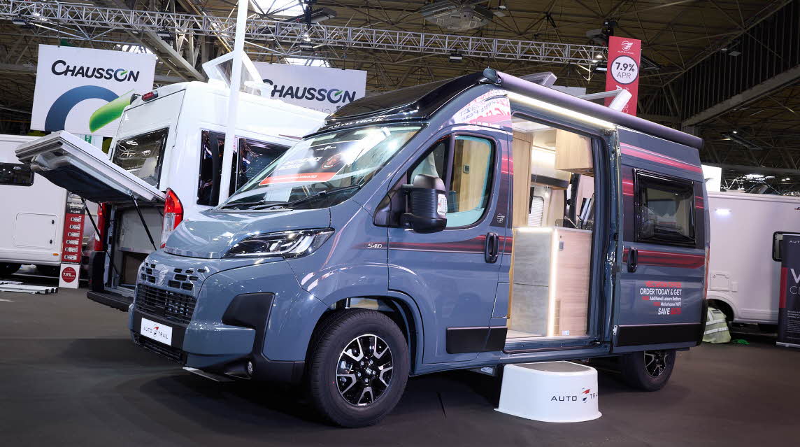 The Auto-Trail V-Line 540 SE is grey with red decals.  There is a show offer on its side in red.  There is a white step by the open sliding door which reveals the interior.  