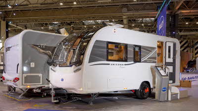 The Bailey Unicorn Deluxe Seville is white with a grey patterned decal.  The front panoramic window is open as is its entrance door.  There is an interactive stand next to two metal steps.