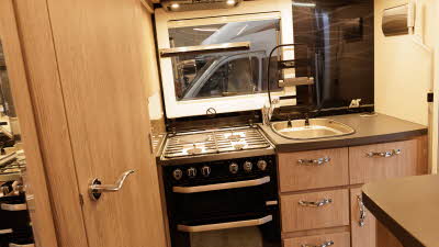 The Auto-Sleeper Nuevo EK Plus kitchen has a full size domestic oven with a square sink.  Both glass lids to the hob and side are open.  The storage cupboards are wooden with large silver handles.