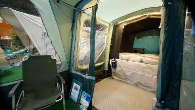 Campmaster 600 Limited edition’s bedroom has a zipped door which is open revealing the bed. There is LED strip lighting on the roof. There is an indoor living area just outside the room with a beige groundsheet. There's a green camping chair.
