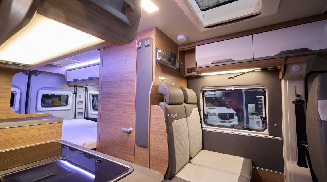 The Knaus Boxlife 600 ME Platinum Selection has white and wooden cabinets.  The upholstery is cream and mid brown leather.  There is a sky light above the sofa.  There is a hob and sink unit in the front, with a fixed rear bed.