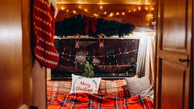 An image framed by open doors with a Christmas stocking on the door, leading into a bed and a large window behind. The bed has a Merry Christmas pillow and fairy lights all around