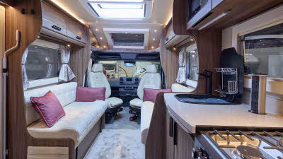 The Auto-Sleeper Burford has a pale grey carpet, with dark wood cabinets.  There are three burgundy cushions.  There is a gas hob with a tower of sockets nearby.  The driver and passenger seats are swivelled and face the interior.