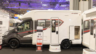 The Auto-Trail F-Line F60 has a grey cab and white coachbuilt body, there are red and grey striped decals on the side.  There is a step in front of the habitation door to gain easy access.  There is an information stand to the left of the step.