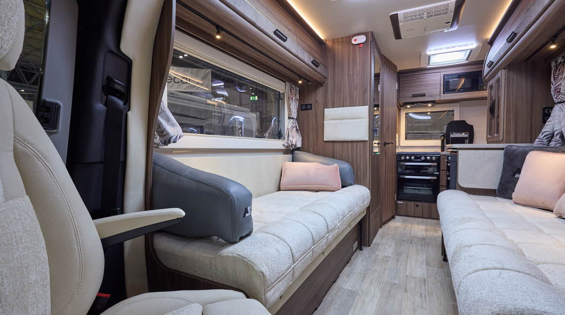 The Auto-Sleeper Bourton has a wooden floor, with dark wood cabinets.  The upholstery is cream with a matching leather surround.  The arms of the sofas are grey and there are grey and peach cushions. The rear kitchen has a black oven.