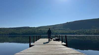 A cleat blue sky above hills and trees in the background with a person walking out onto a pier over a beautiful lake