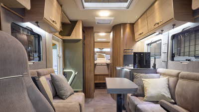 The Eura Mobil Integra Line GT 726 EF has beige carpets and wooden cabinets.  The upholstery is mid brown.  There is a grey table (folded) in front of the sofas.  There are two steps to the rear fixed bed.