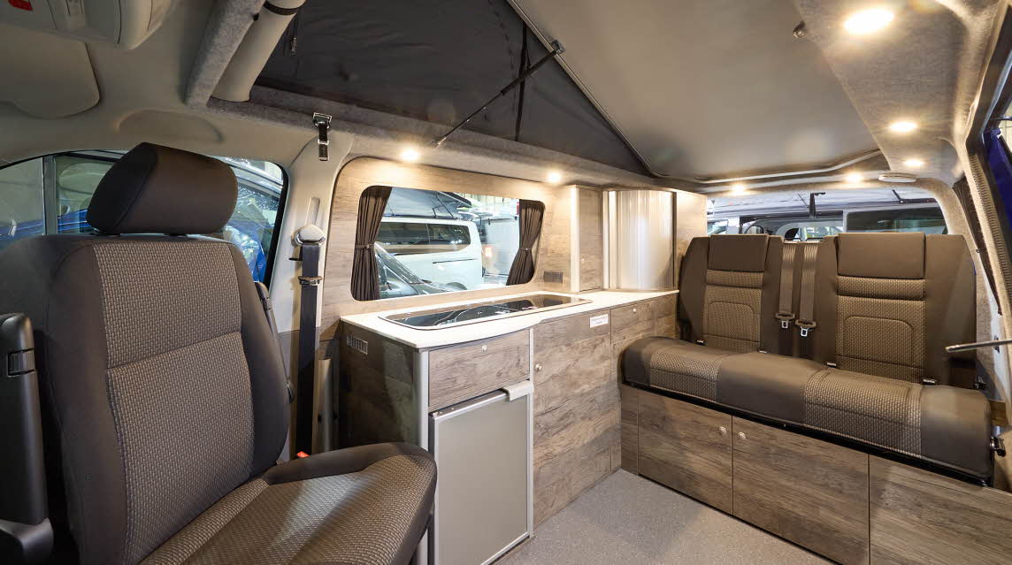 The Rolling Homes Camper Ltd Shackleton has beige flooring.  The upholstery is brown with a matching pattern.  The cabinets are wooden.  The rising roof is fully extended.