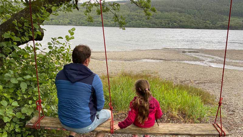 Dad and daughter sitting on a swing looking across at a lake