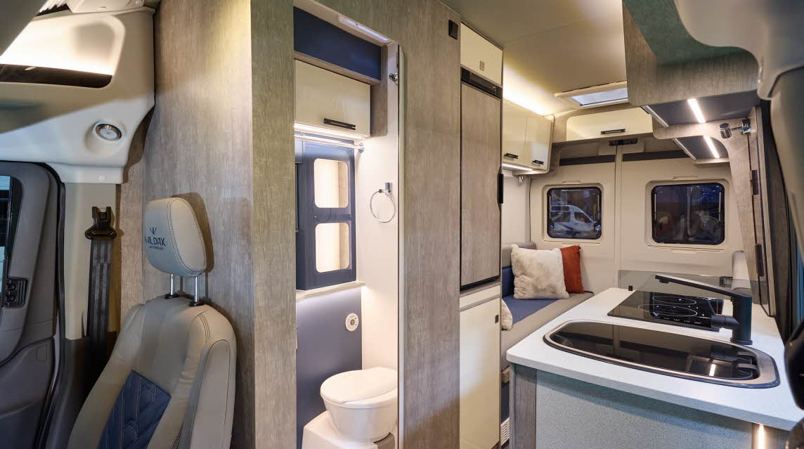 WildAx Motorhomes Meteor has wooden cabinets, its upholstery is pale leather with a blue inset.  The washroom door is open revealing the toilet.  There is a rear lounge.