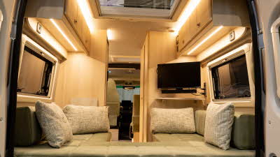 The Vantage TEO has green upholstery and a u shaped sofa.  It has pale wood cabinets.  There is a cream cushion in each corner of the sofa.  There is TV in the right corner and a large skylight