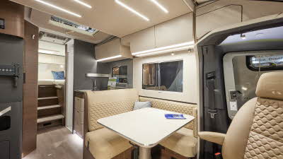 Adria Matrix Supreme MB 670 SL has cream embossed upholstery, with cream overhead cabinets and wooden curved panelling, it has mid-brown wooden flooring, there are three steps leading to the fixed rear bed with a blue cushion with lots of lighting.