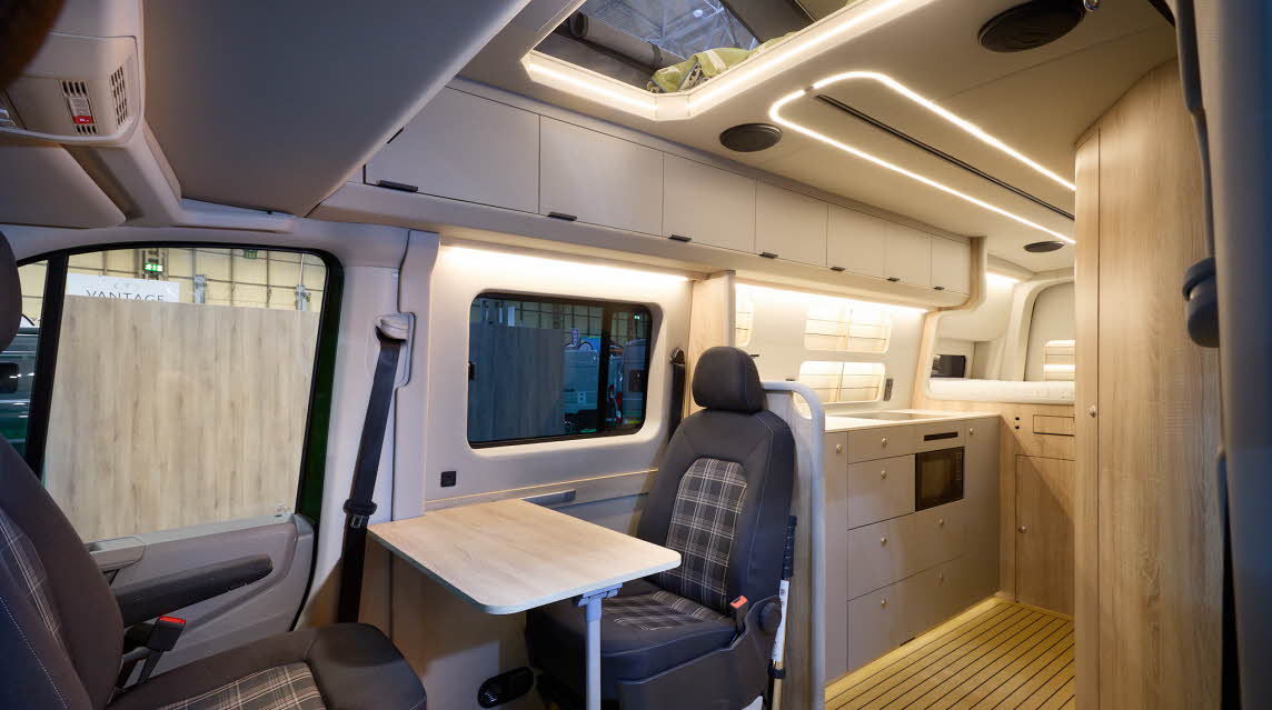 The Kaktus Adventure Vans Oasis x4 has a wooden floor.  The upholstery is two tone brown with a tartan pattern.  The cabinets are all pale wood.  The pop top roof is extended.