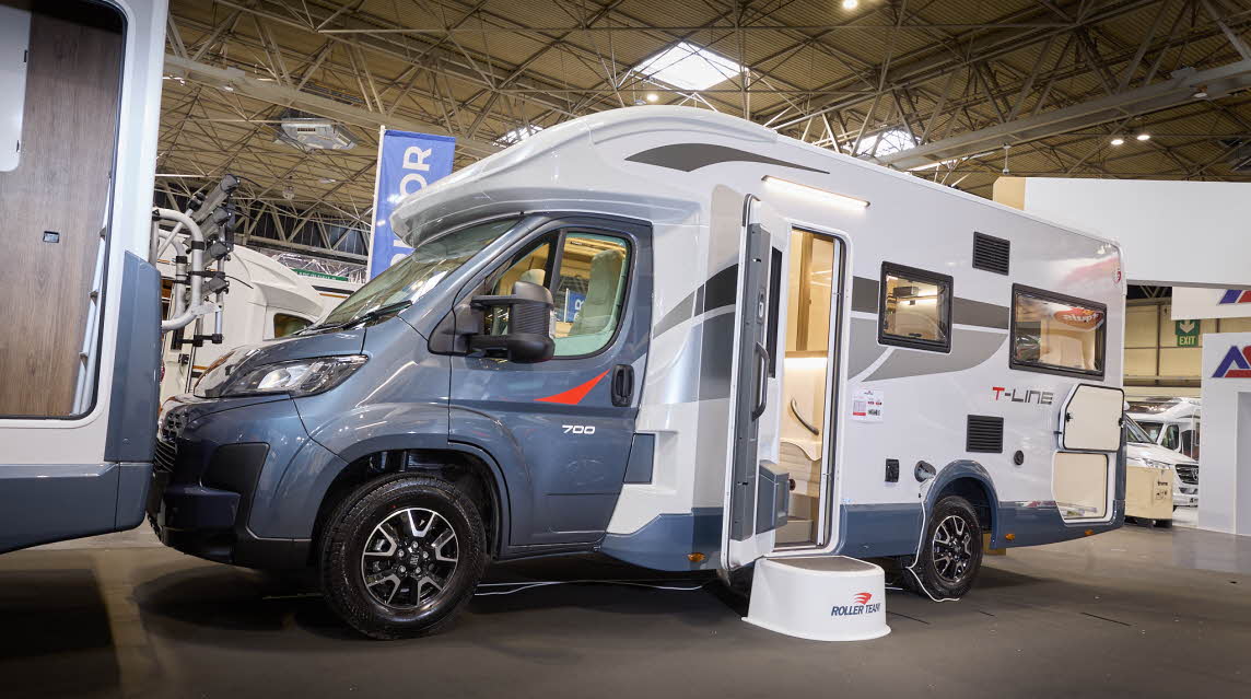 The Roller Team T Line 700 has a blue cab and its body is white with blue trim with grey and red decals.  There is a white step outside the open habitation door.  The rear locker door is open.