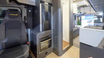 The Swift Trekker XL has a wooden floor with a step up into the grey carpet at the rear. There is an oven behind the driver's seat. The washroom door is open. There is a rear lounge.