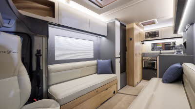 The Auto-Trail Excel 620S has beige carpet, with pale wood cabinets with white overhead lockers. The upholstery is cream leather. There are blue cushions. The kitchen is at the end with an oven and a microwave in an overhead locker.