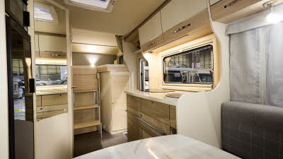 Eura Mobil Activa One’s interior has dark grey upholstery.  Its furniture is a pale wood with cream doors.  There is a high fixed bed at the rear with a ladder for easy access.  Underneath the bed there is a wardrobe.