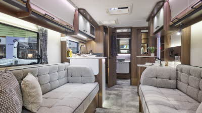 Interior of the Coachman Laser 480 Xtra with grey sofas, cosy cushions and wood accents and parquet effect flooring.  The door to the rear washroom is open.  Perfect for relaxation and designed for comfort.