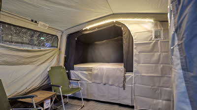 In the Campmaster 1000 LX there is a coffe table and chairs. The bedroom door is open. There is LED strip lighting on the ceiling.