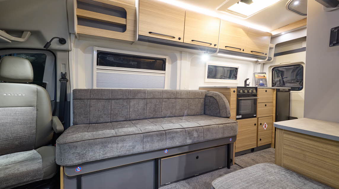 The Bailey Endeavour B63 has grey carpets, with wooden and grey cabinets.  The upholstery is also grey.  There is a kitchen to the rear of the van.  