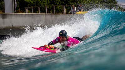 A surfer wearing a helmet laying flat on the surfboard on a wave of blue and white as the wave breaks, with a wall in the background