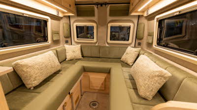 The Vantage NEO has a green u shaped sofa with patterned cushions.  The cabinets are made of pale wood and it has removable carpets on the floor.