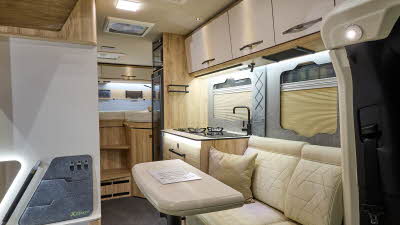 Eura Mobil Xtura XT’s interior has cream upholstery.  Its furniture is pale wood with cream doors.  The table can be extended and is showing folded in half.  At the rear there are two fixed beds.  