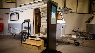 The classically styled Eriba Touring 420 Legend has a two tone layout in silver and white.  Its pop top roof is open as is its entrance door.  There are two wooden steps to gain easy access.